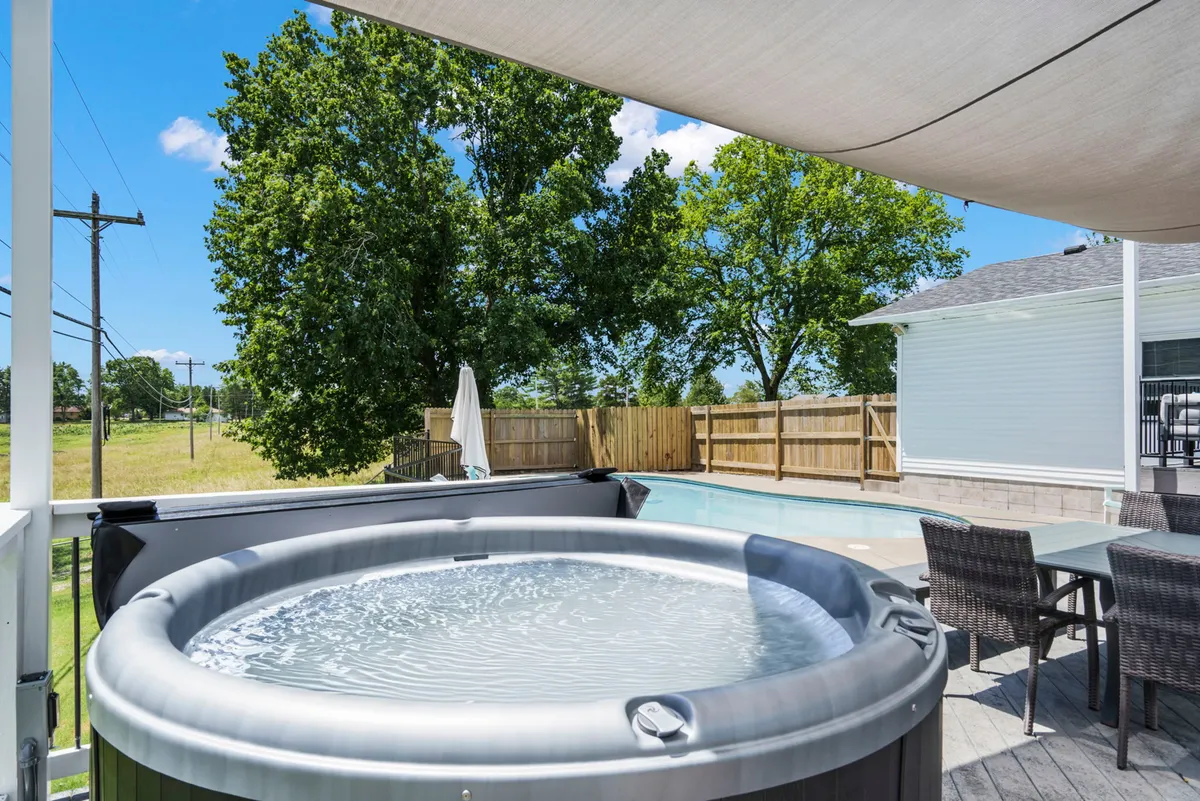 This exterior image features a hot tub and swimming pool set amidst leafy green trees. A spacious outdoor dining area provides a welcoming spot for group gatherings or meals under the sky. The scene captures friends relaxing in the hot tub and others mingling around the pool, with the landscape evoking an inviting, social atmosphere.