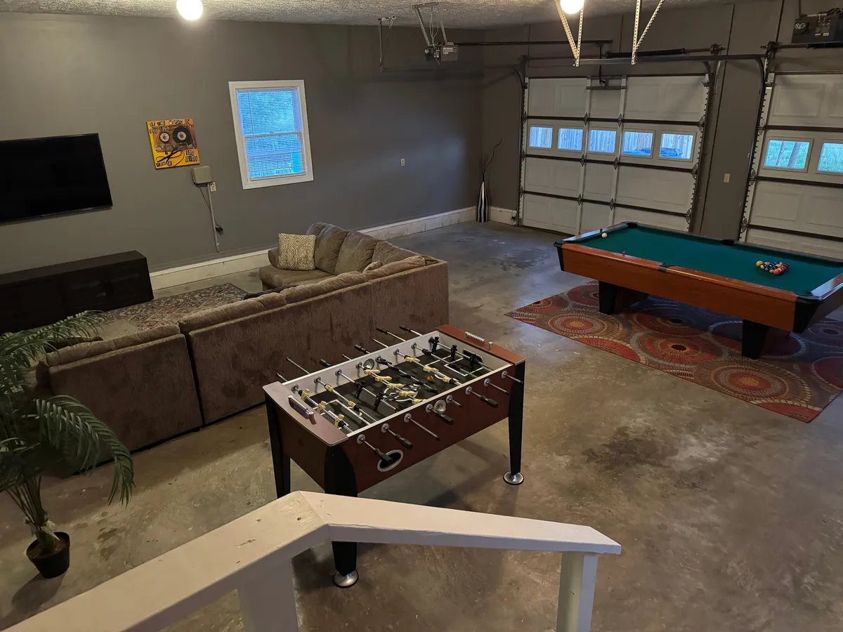 A spacious, vibrant garage game room featuring a foosball table and a pool table as the central focus. A cozy sofa sits nearby, perfect for relaxing or watching others compete. The space is decorated with playful wall art and has plenty of room for group activities.
