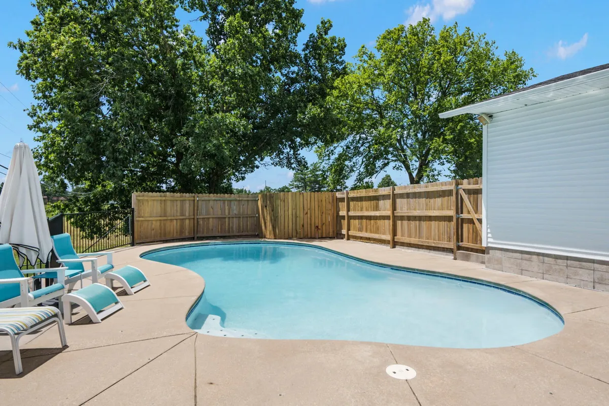 This exterior image displays a spacious backyard featuring a well-maintained swimming pool, lounge chairs for relaxation, and, notably, a fenced-in green lawn bordered by mature trees. The fencing is clearly visible, suggesting a secure outdoor area ideal for both human and canine playtime. The relaxed atmosphere is enhanced by the tranquil pool and surrounding natural elements.
